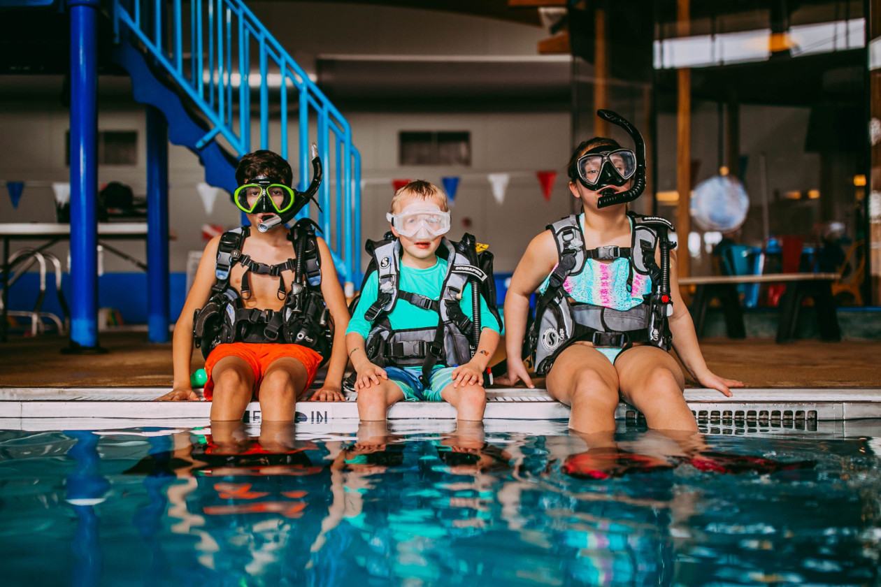 three kids in scuba gear sitting on a diving platform