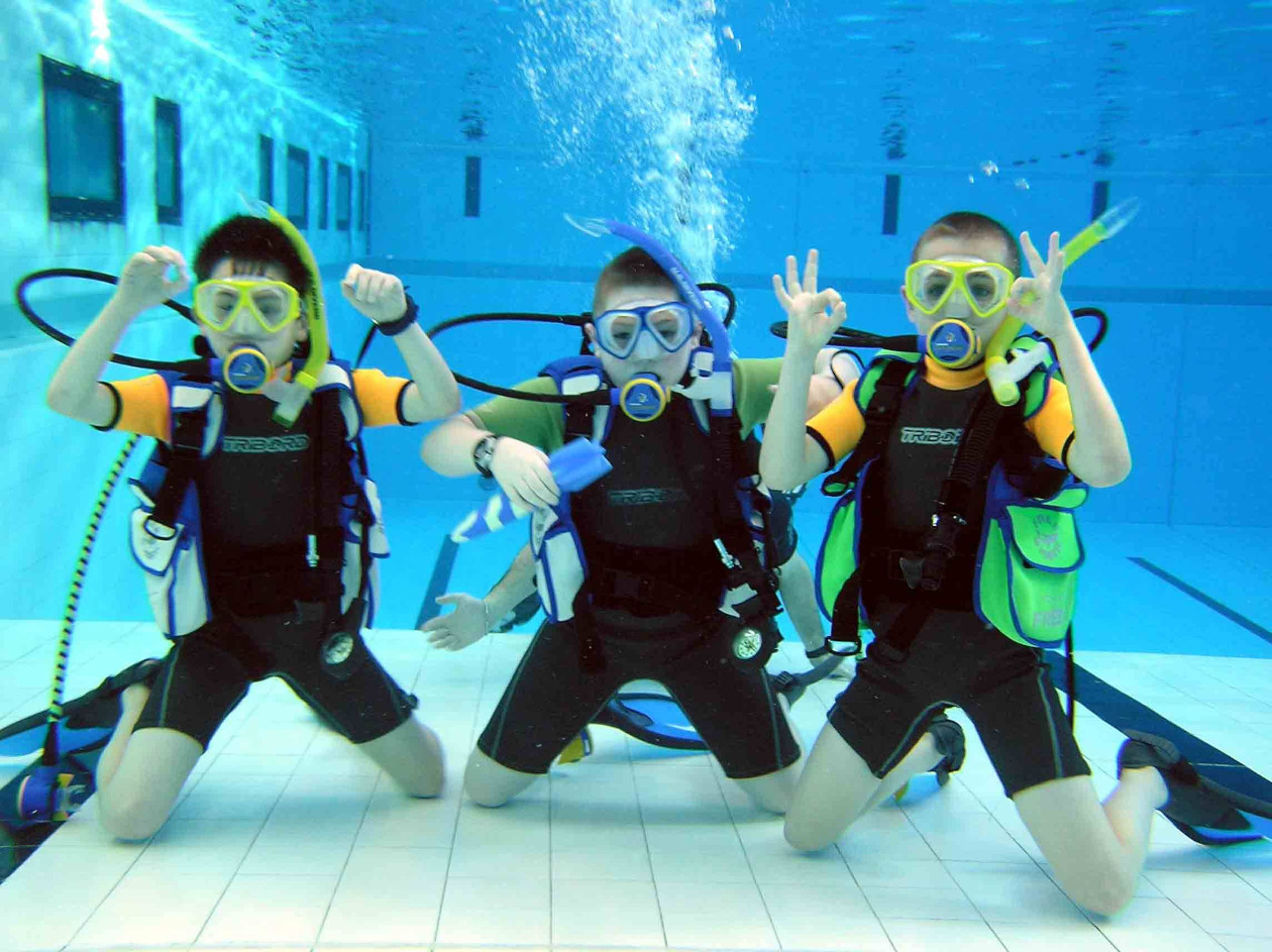 Tres niños practicando buceo en una piscina, mientras hacen señas con las manos